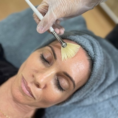 Mulher com toalha cinzenta na cabeça recebe aplicação facial com pincel branco