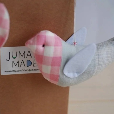 Pássaro decorativo em tecido com padrão xadrez rosa e branco e asas brancas, com etiqueta JUMA MADE
