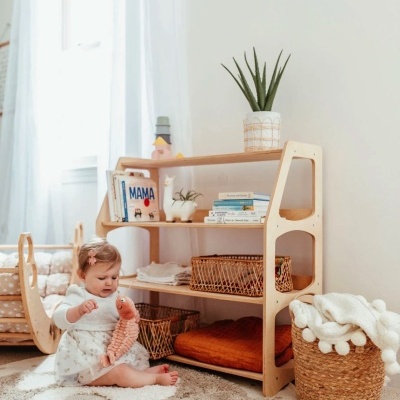 Estante de madeira com livros, cestos e planta num quarto claro com criança sentada no tapete