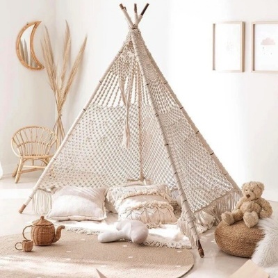 Tenda tipi de croché bege em ambiente claro com almofadas, baloiço de madeira, cadeira de vime e urso de peluche.