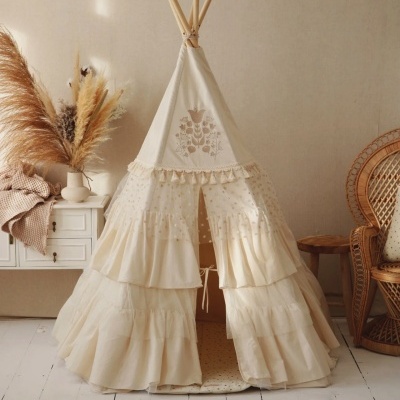 Tenda tipi infantil bege com varas de madeira e decoração ao lado em ambiente claro