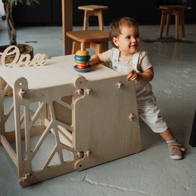 Criança brinca com toy de madeira numa mesa infantil em interior com chão cinzento e bancos de madeira