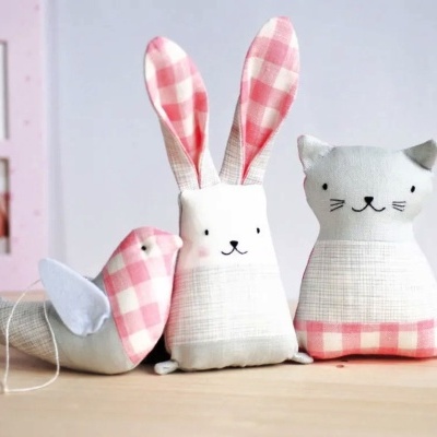 Três brinquedos de pelúcia representando animal com padrão xadrez rosa sobre madeira, fundo branco com quadros na parede