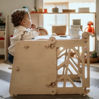 Criança junto a mesa de madeira clara com recortes geométricos em ambiente interior com brinquedos ao fundo