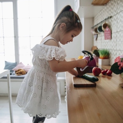 Menina de vestido branco a lavar prato numa cozinha com bancada de madeira e frutas ao lado