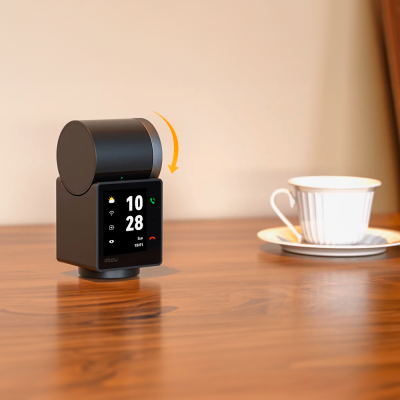 Câmara de vigilância preta com ecrã digital sobre mesa de madeira e chávena ao fundo