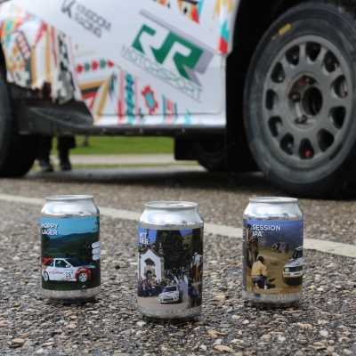 Três latas de bebida no chão com carro branco decorado ao fundo