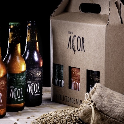 Três garrafas de cerveja ACOR ao lado de caixa de embalagem e saco com grãos