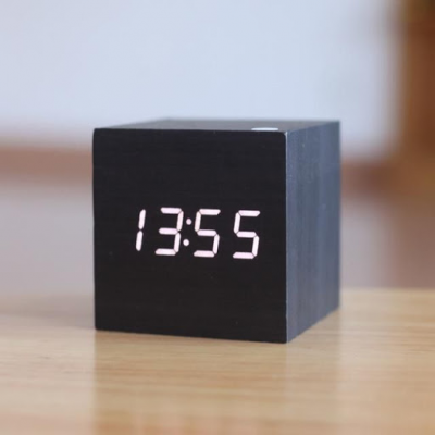 Relógio digital preto em formato cubo com horas 13:55 sentado numa mesa de madeira
