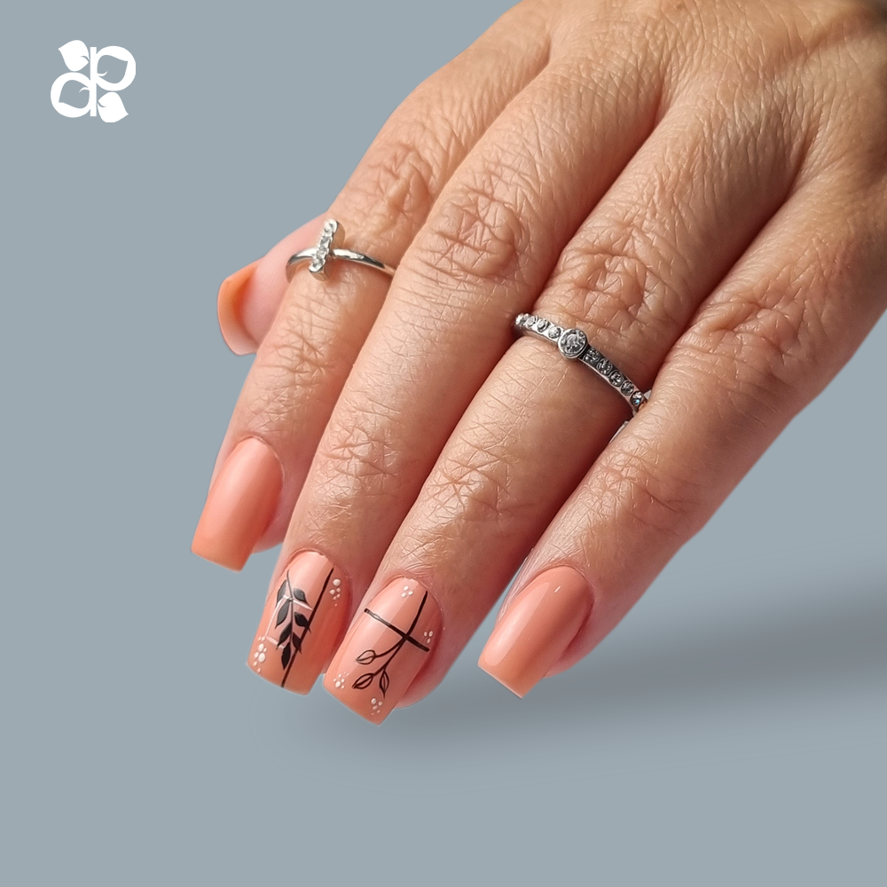 Mão feminina com unhas pintadas de coral e desenhos de folhas pretas, com dois anéis prateados, fundo cinza.