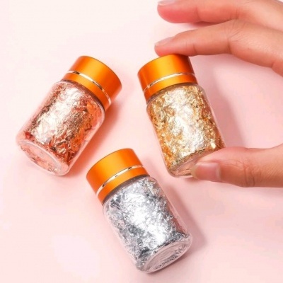 Três frascos com folhas metálicas decorativas em cores cobre, dourado e prateado com tampas laranja, sobre fundo rosa