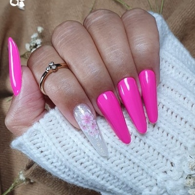 Mão com unhas longas rosa fluorescente e branca com flor, anel dourado, fundo tecido castanho e malha branca.