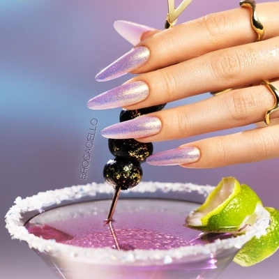 Unhas com verniz gel cintilante rosa com pedras pretas decorativas, copo de cocktail com borda salgada e lima numa mesa com fundo rosa e azul