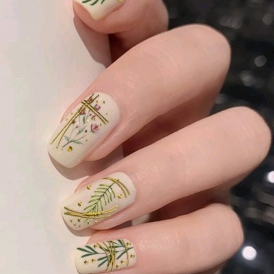 Unhas pintadas com verniz creme e decoração de folhas verdes, linhas douradas e pequenos detalhes florais