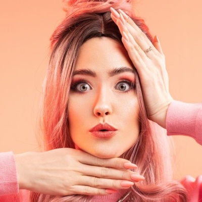 Mulher com cabelo rosa e maquiagem, camisola rosa, fundo laranja