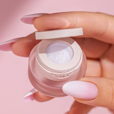 Frasco de vidro fosco rosado com tampa branca, segurado por mãos com unhas compridas e esmalte rosa claro