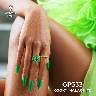 Anel verde e dourado com cristal roxo em mão com unhas verdes sobre fundo e roupa verde