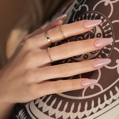 Mão feminina com unhas rosa nude e anéis dourados sobre tecido padrão geométrico castanho e branco.