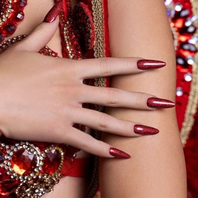 Mão feminina com unhas vermelhas brilhantes sobre braço e vestido com pedras vermelhas e detalhes dourados