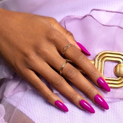 Mão com unhas rosa vibrante, anéis dourados e roupa lilás com cinto dourado