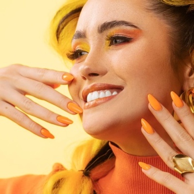 Mulher sorridente com maquilhagem e unhas laranja, acessórios dourados