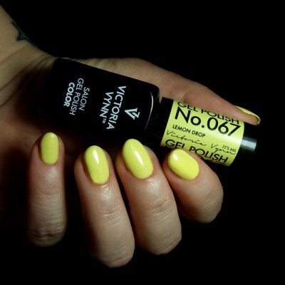 Mãos femininas com unhas pintadas de verniz gel amarelo segurando dois frascos de verniz gel, um preto e um amarelo.