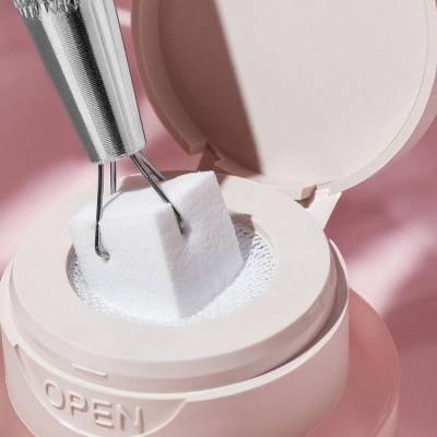 Estojo de pó compacto branco com tampa aberta e cubo branco a ser segurado com pinça.