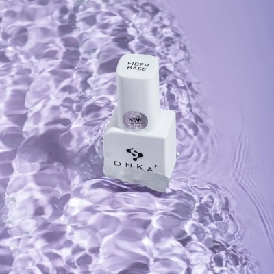 Frasco branco de verniz para unhas DNKA' sobre superfície lilás com líquido translúcido