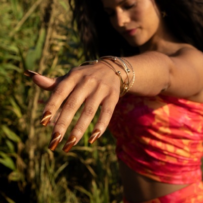 Mulher com pulseiras douradas, unhas castanho metálico e top rosa e laranja ao ar livre