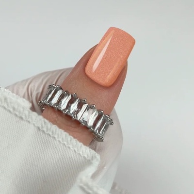 Anel prateado com pedras transparentes em dedo com unha pêssego brilhante e manga branca.