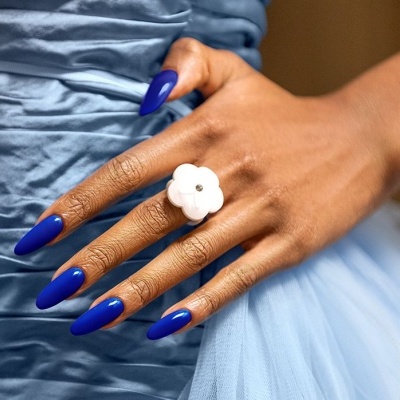Mão com unhas azuis e anel de flor branco sobre vestido azul