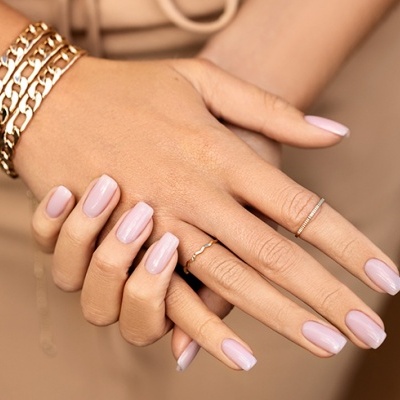 Mãos femininas com unhas rosa claro fosco, anéis e pulseiras dourados.