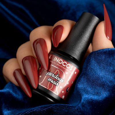 Frasco de verniz INOCOS em mão com unhas vermelhas, fundo azul brilhante