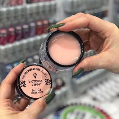 Frasco de gel para unhas Victoria Vynn com creme cor de pele segurado por mãos com unhas verdes