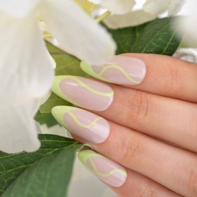 Unhas com design verde claro ondulado sobre base transparente, entre flores e folhas brancas.