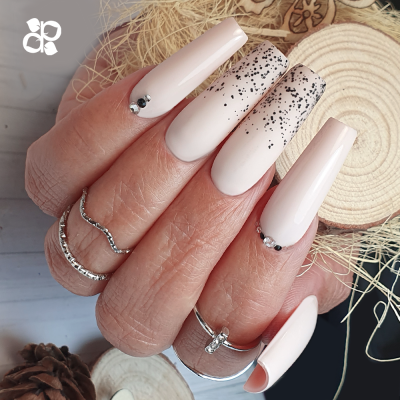 Unhas longas decoradas com esmalte rosa claro, cristais e glitter, com anéis prateados na mão em fundo rústico.