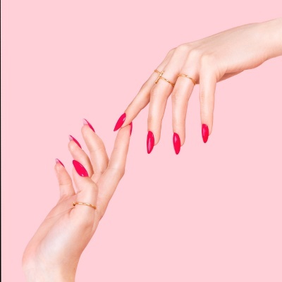 Mãos femininas com unhas vermelhas e anéis dourados sobre fundo rosa
