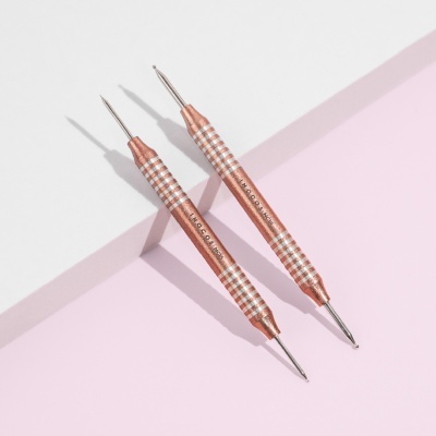 Dois instrumentos metálicos cobre com padrão xadrez branco, pontas duplas, sobre fundo branco e rosa