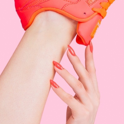 Ténis desportivo laranja vibrante com mão feminina e unhas cor de laranja sobre fundo rosa