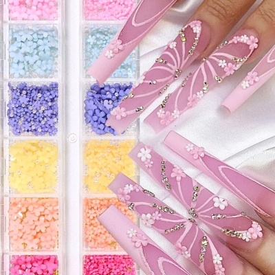 Unhas postiças rosas decoradas com flores 3D brancas e pedras, ao lado de caixa com flores decorativas coloridas.