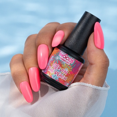 Frasco de verniz gel INOCOS cor rosa em mão com unhas rosa junto a fundo azul água