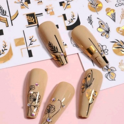 Unhas postiças bege decoradas com padrões florais e geométricos em preto e dourado, com pequenos strass.