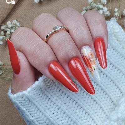 Mão feminina com unhas vermelhas compridas e decoração floral numa unha, com anel dourado e roupa de malha branca.