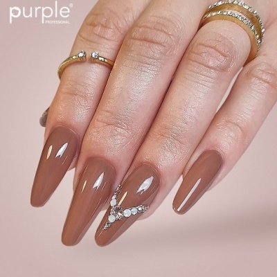 Mano femenina con uñas marrón brillante decoradas con pedrería y anillos dorados sobre fondo rosa