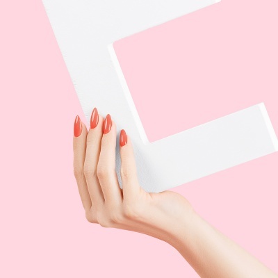 Mão feminina com unhas vermelhas segurando letra E branca sobre fundo rosa