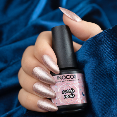 Mão com unhas compridas pintadas segurando frasco de verniz Inocos 'sorrir mais' sobre fundo azul.