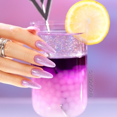Unhas com verniz gel lilás glitter segurando copo com bebida rosa e roxa e rodela de limão