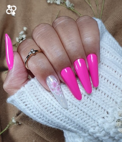 Mão com unhas longas rosa fluorescente e branca com flor, anel dourado, fundo tecido castanho e malha branca.