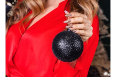 Bola de Natal preta brilhante sendo segurada por mão com unhas douradas, vestido vermelho ao fundo