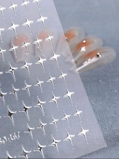 Folha de adesivos prateados em forma de estrelas e unhas artificiais translúcidas com detalhes laranja.